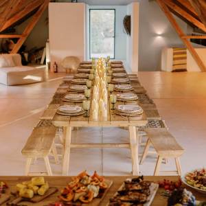 Long dining table in rustic room set with candles and plates, food spread in foreground.