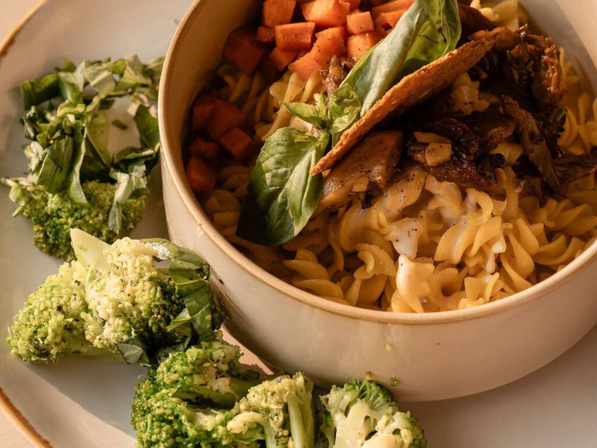 Bowl with pasta, carrots, mushrooms, and basil, served on a plate with broccoli and herbs.
