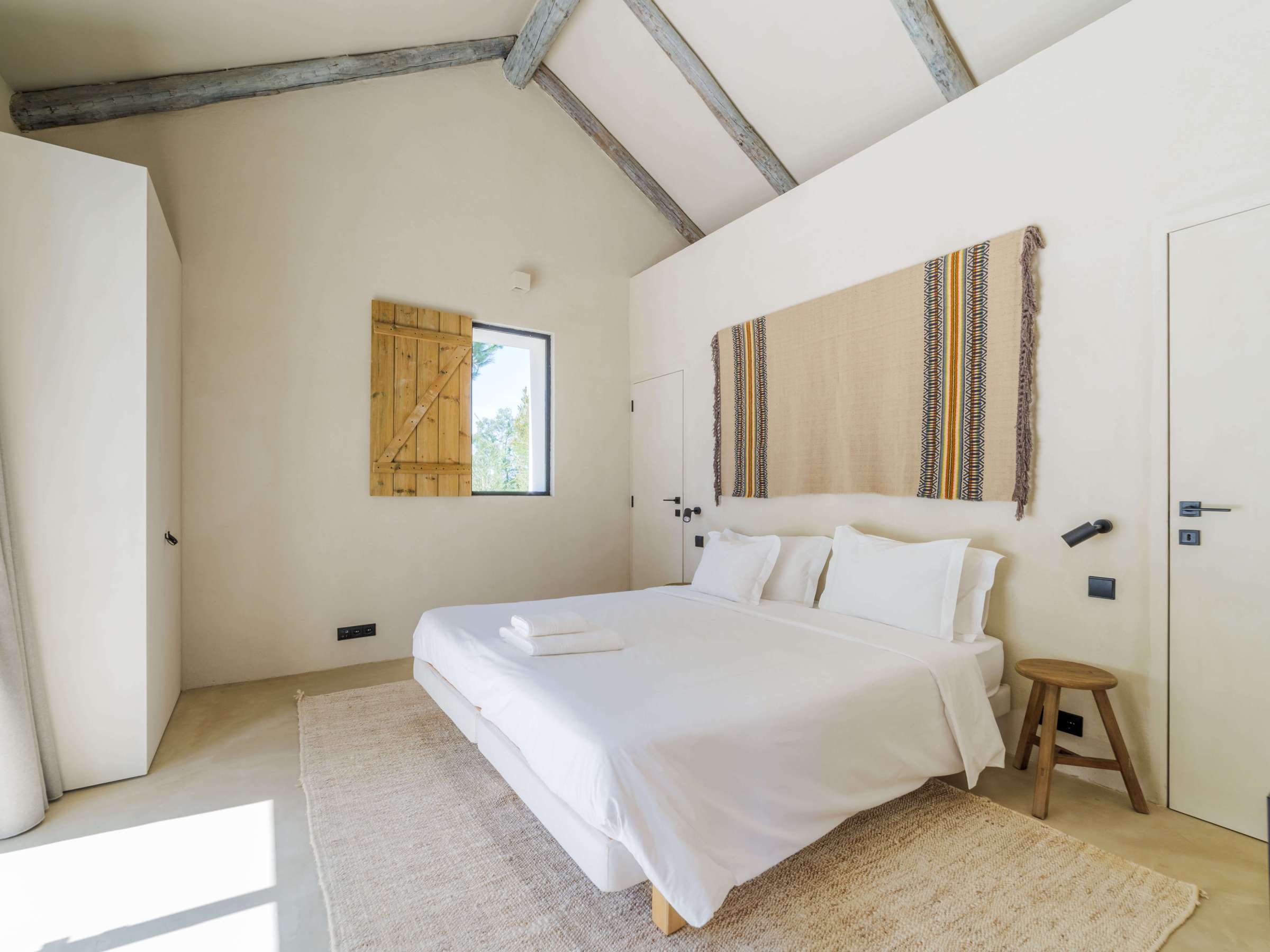 Minimalist bedroom with double bed, wooden stool, and woven wall art.