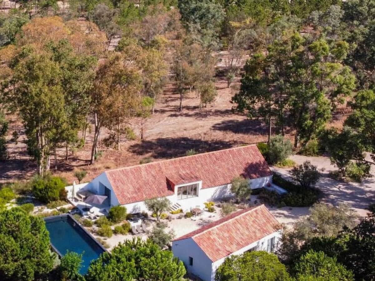 Aerial view of a house with white walls, a red roof, pool, and surrounding trees in a rural area.