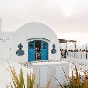 White domed building with blue doors, chairs visible inside, outdoor tables on the right, plants in foreground.